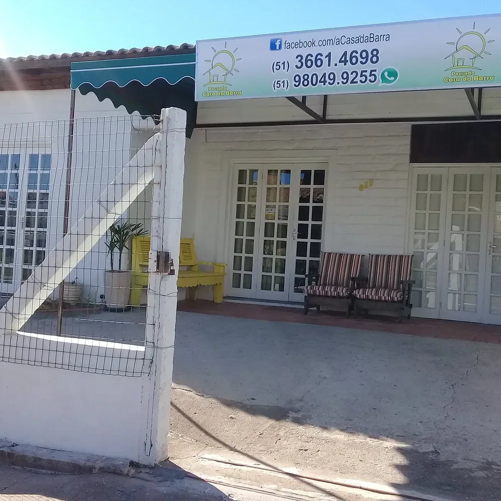 Entrada da Pousada Casa da Barra de dia, com banco amarelo, poltronas listradas na varanda e placa com telefone e WhatsApp
